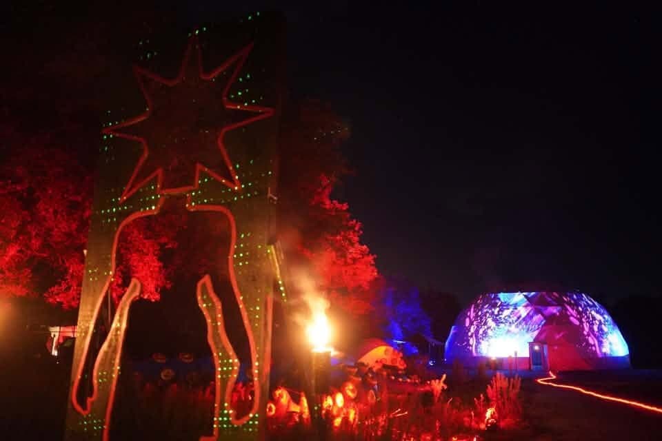 A towering figure with a star-shaped head, outlined in red lights, stands beside a fire, with the projection-lit dome beyond